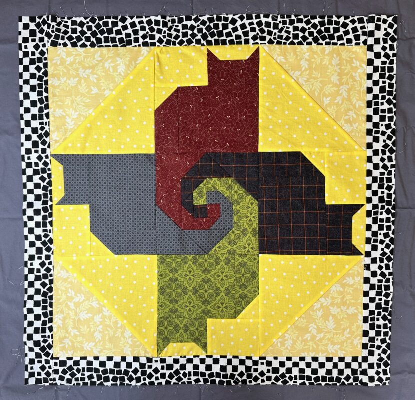 Small, 2 foot square quilt top showing 4 cats with their tails wrapped around each other in the middle. Yellow polka dot fabric separates the cats. There is a black and white, chunky, border.