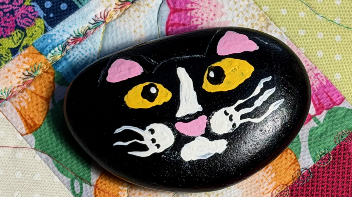 Smiling black cat with yellow eyes and pink ears painted on a rock. Rock Painting