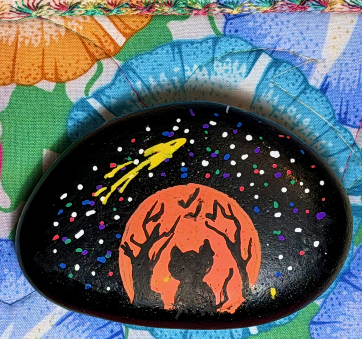 Painted rock showing black cat and trees against an orange moon, with stars, including a falling star, shown above. Rock Painting
